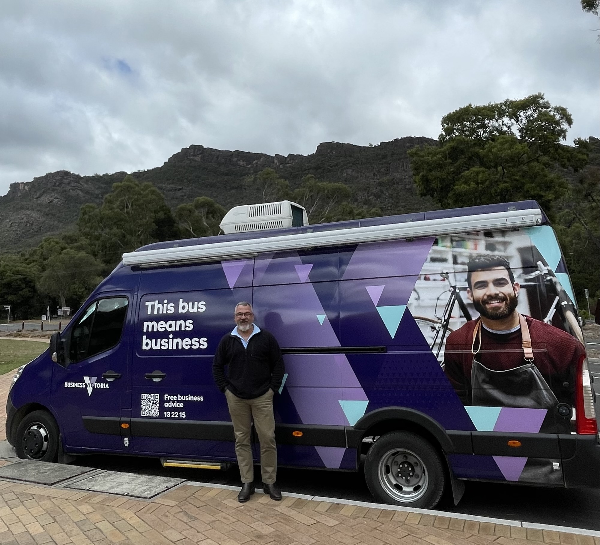 A man standing in front of a purple bus, posing, with an image of a man in a red shirt to the right. The sky is cloudy.